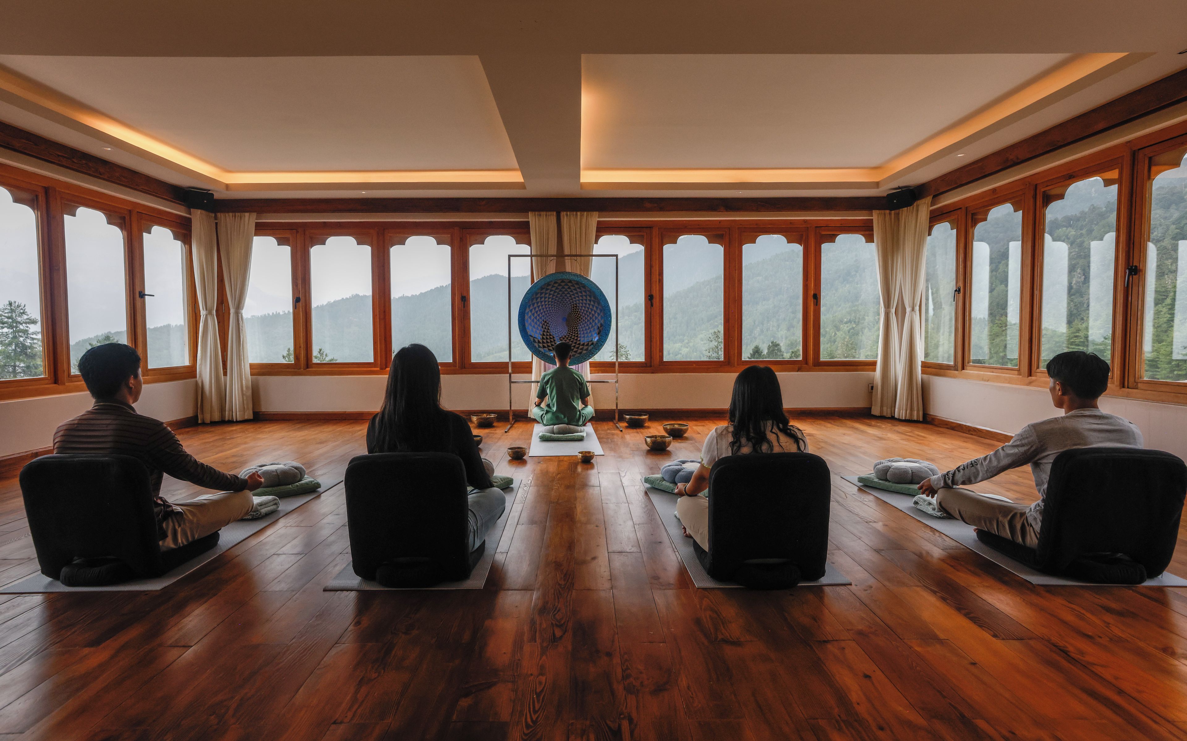 A room with wooden floors, large windows, and a central meditation area with cushions and bowls.