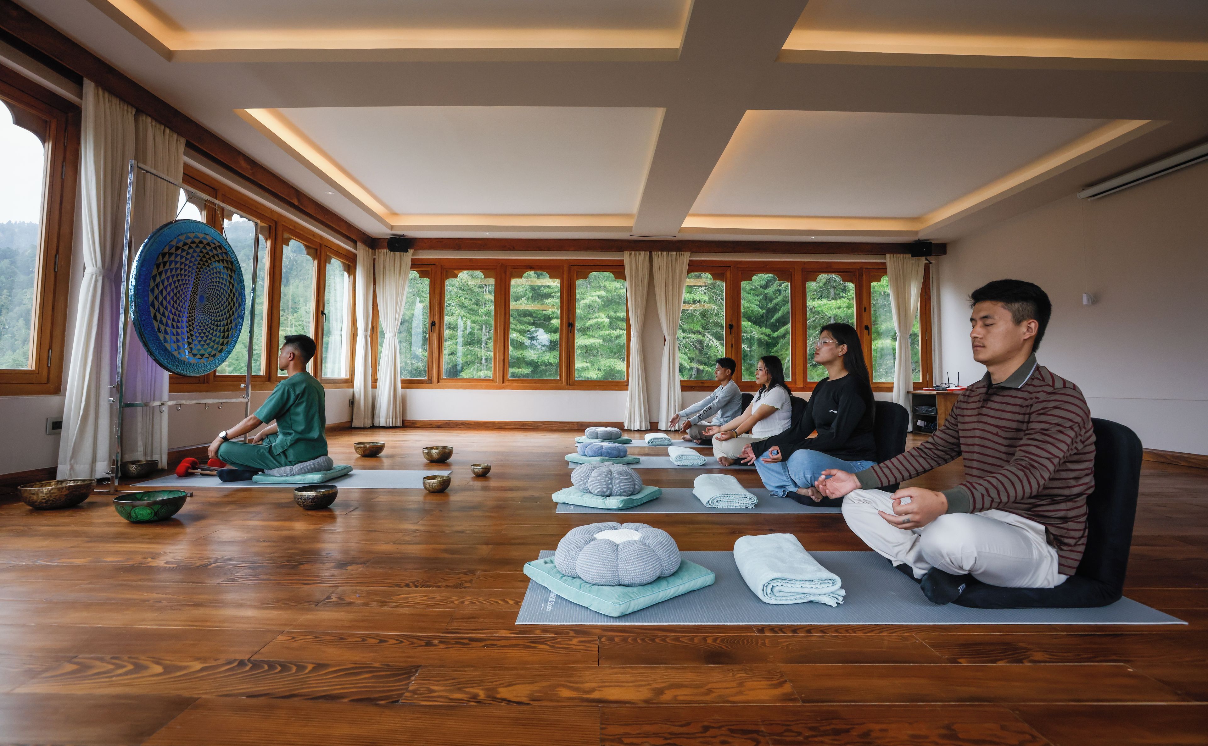 Clase de yoga en una habitación con suelo de madera, ventanas amplias y vistas a la vegetación.