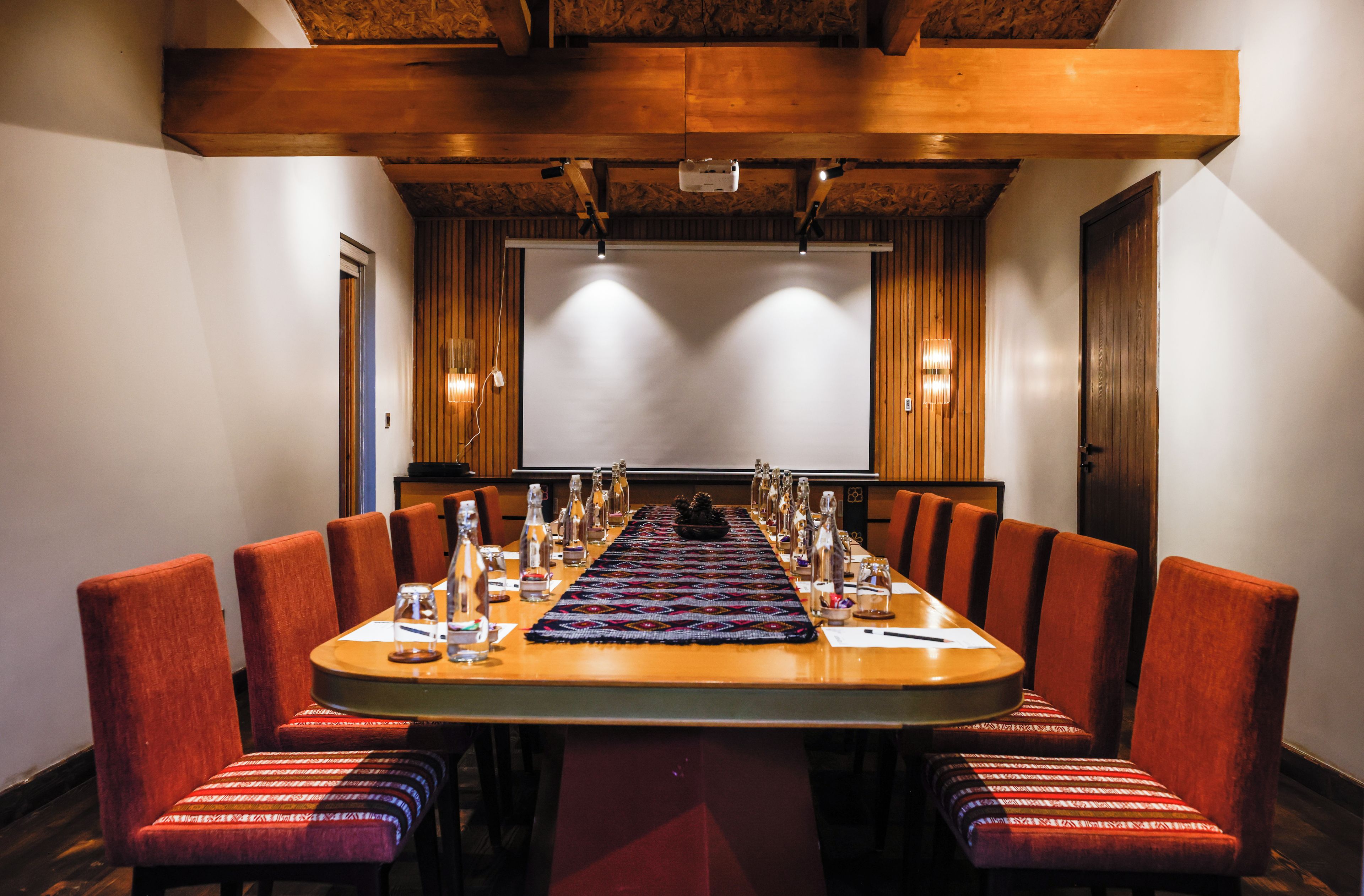 A conference room with a long table, red upholstered chairs, and a projector screen.