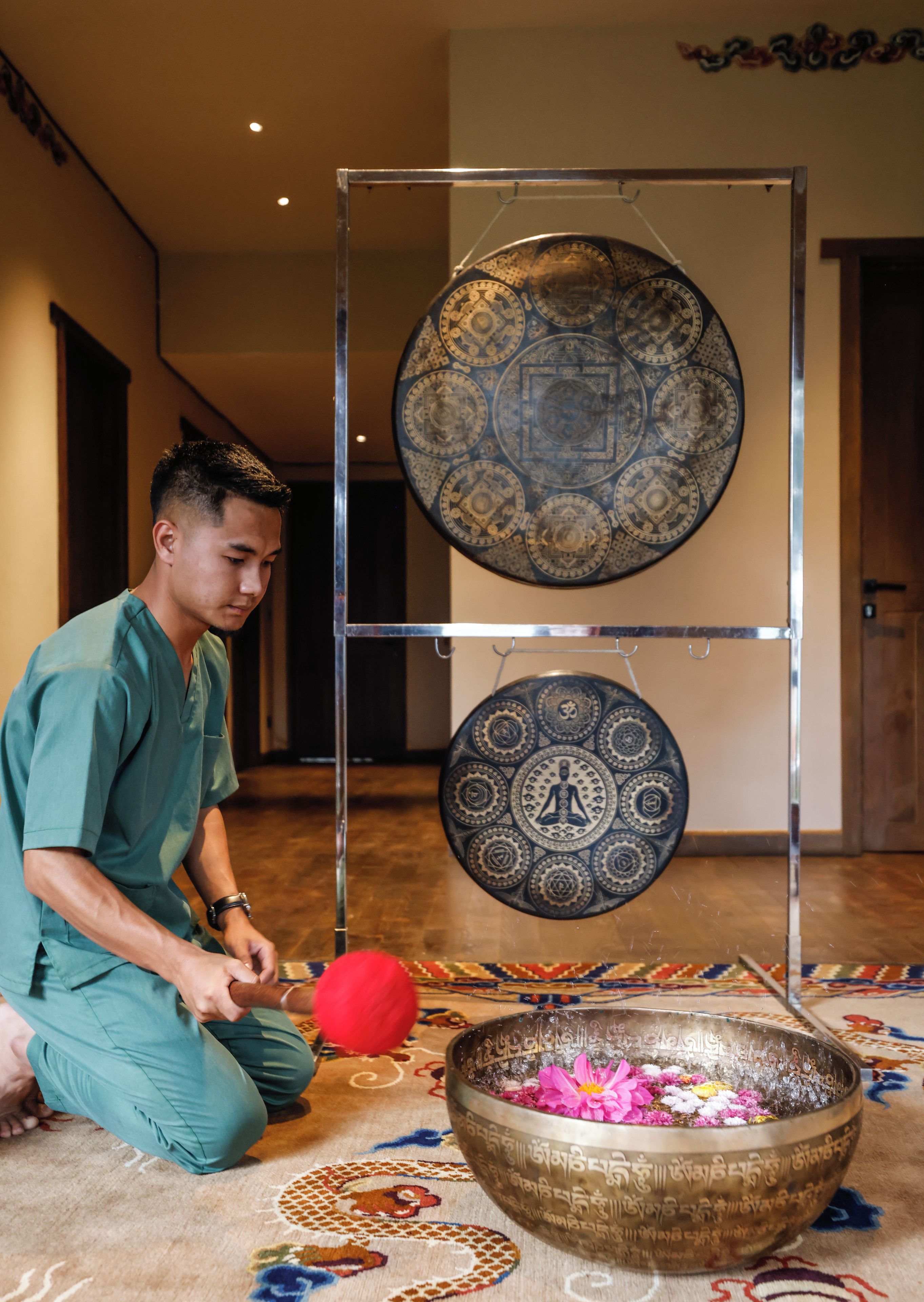 A person in medical scrubs kneeling on a patterned carpet with a large brass bowl containing flowers and a red ball.