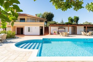 Una casa de dos pisos con piscina, mobiliario exterior y un patio cubierto.