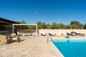Un área junto a la piscina con sillones de descanso, un espacio de estar cubierto y un patio de piedra.