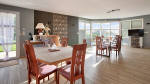 Un comedor amplio con una mesa grande y varias sillas, una cocina con televisor y chimenea, y ventanas grandes que ofrecen vistas al exterior.