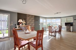 Un vaste espace de salle à manger avec une grande table et de nombreuses chaises, un coin cuisine équipé d’un téléviseur et d’une cheminée, ainsi que de grandes fenêtres offrant une vue sur l’extérieur.