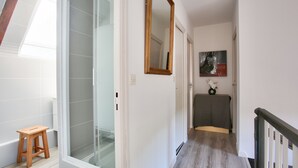 Un pasillo con un taburete de madera, una puerta de ducha de vidrio, un espejo y un cuadro en la pared.