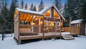 A wooden cabin with a deck and a covered porch, surrounded by snow and trees.