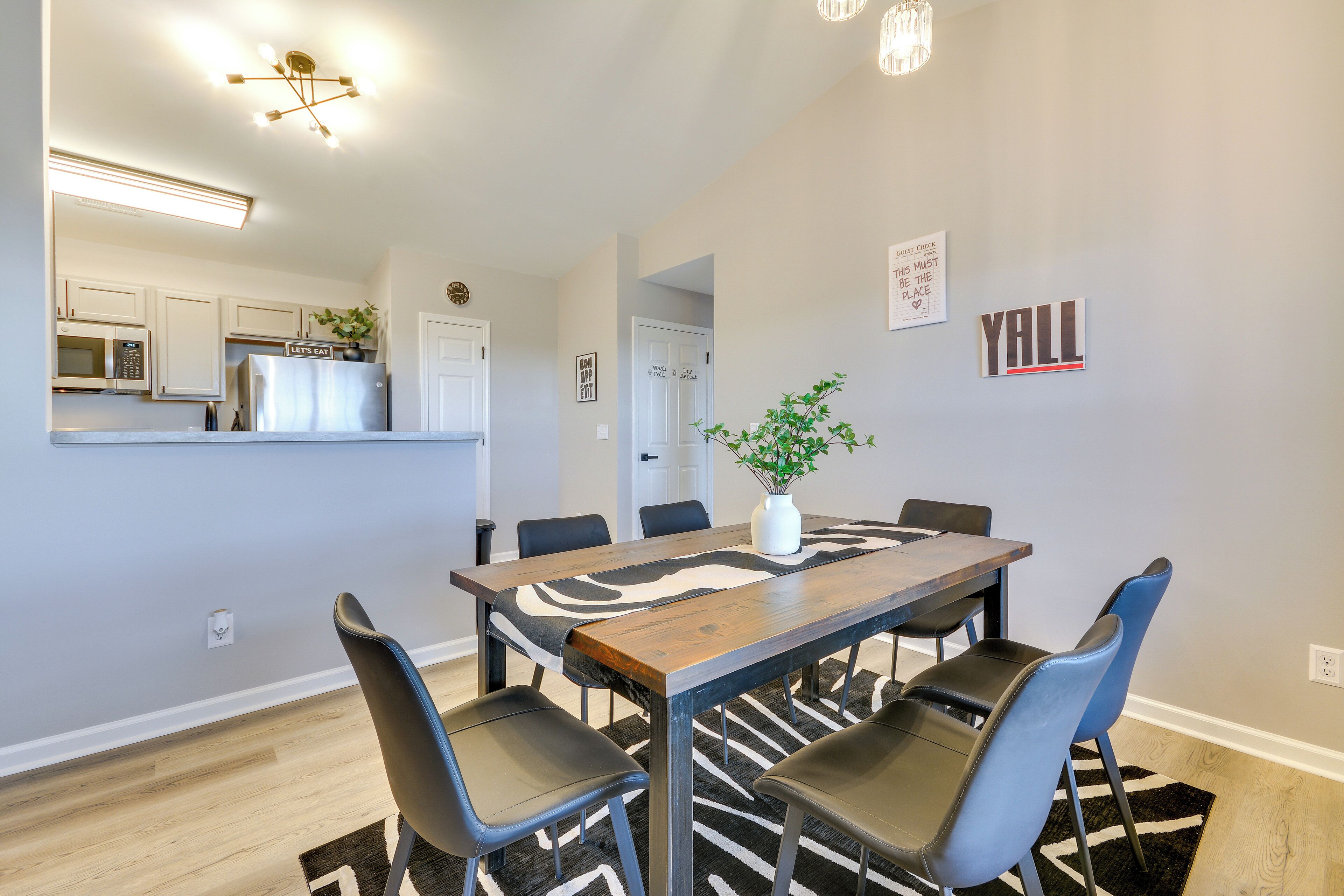 A dining area with a wooden table and six chairs, a kitchen in the background, and a 'Yall' sign on the wall.