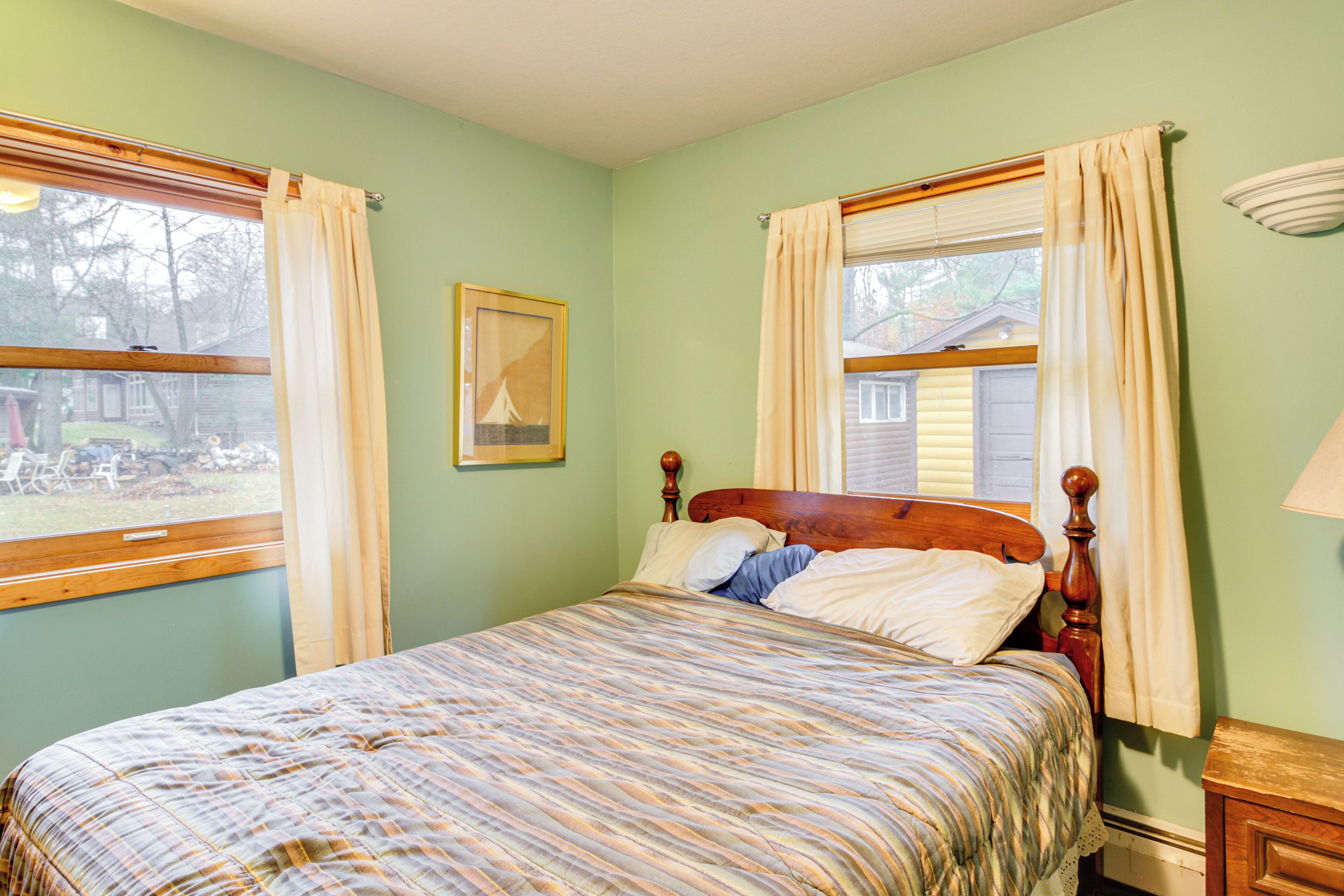 A bedroom with a bed, two windows with curtains, a framed picture on the wall, and a nightstand with a lamp.