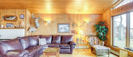 A wooden-paneled living room with a leather sofa, a small table, a wicker chair, and a large window.