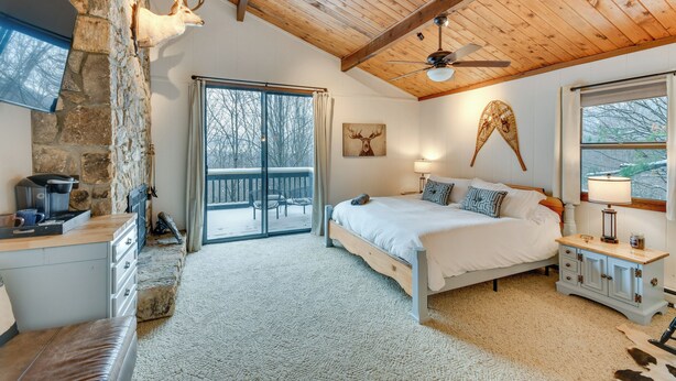 A bedroom with a stone fireplace, a bed with a wooden headboard, a nightstand, and a large window with a view of trees. - 6 Mi to Skiing! Hot Tub Hideaway in Seven Devils (Banner Elk)