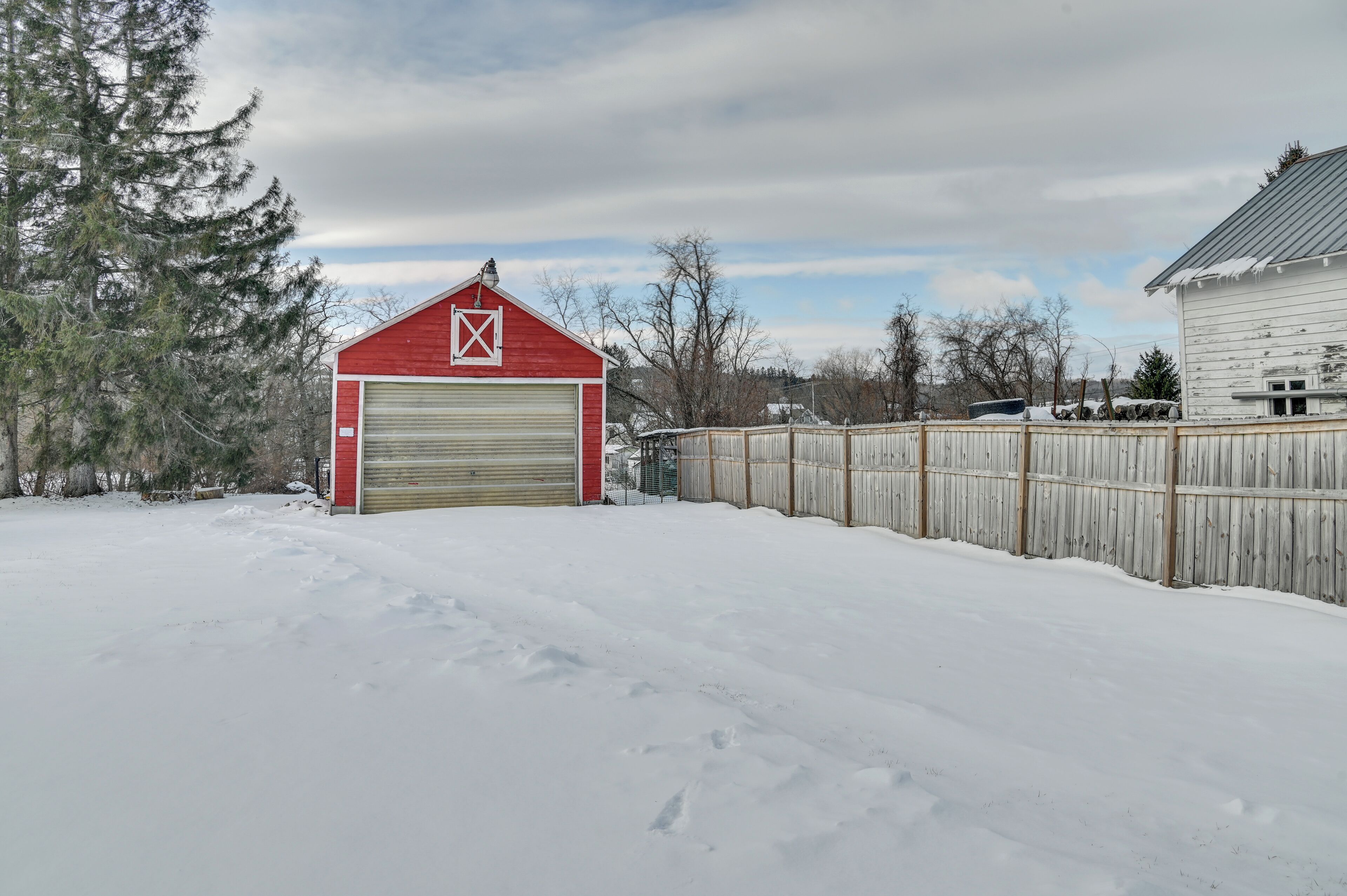 Un garage enneigé, à l’extérieur rouge et à la porte blanche, entouré d’une clôture en bois et d’arbres.