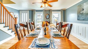A dining room with a wooden table set for six, surrounded by chairs, a ceiling fan, and a large window with curtains.