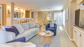 Ein modernes Wohnzimmer mit Sofa, Sessel und Couchtisch. Im Hintergrund ist ein Essbereich mit Tisch und Stühlen zu sehen.