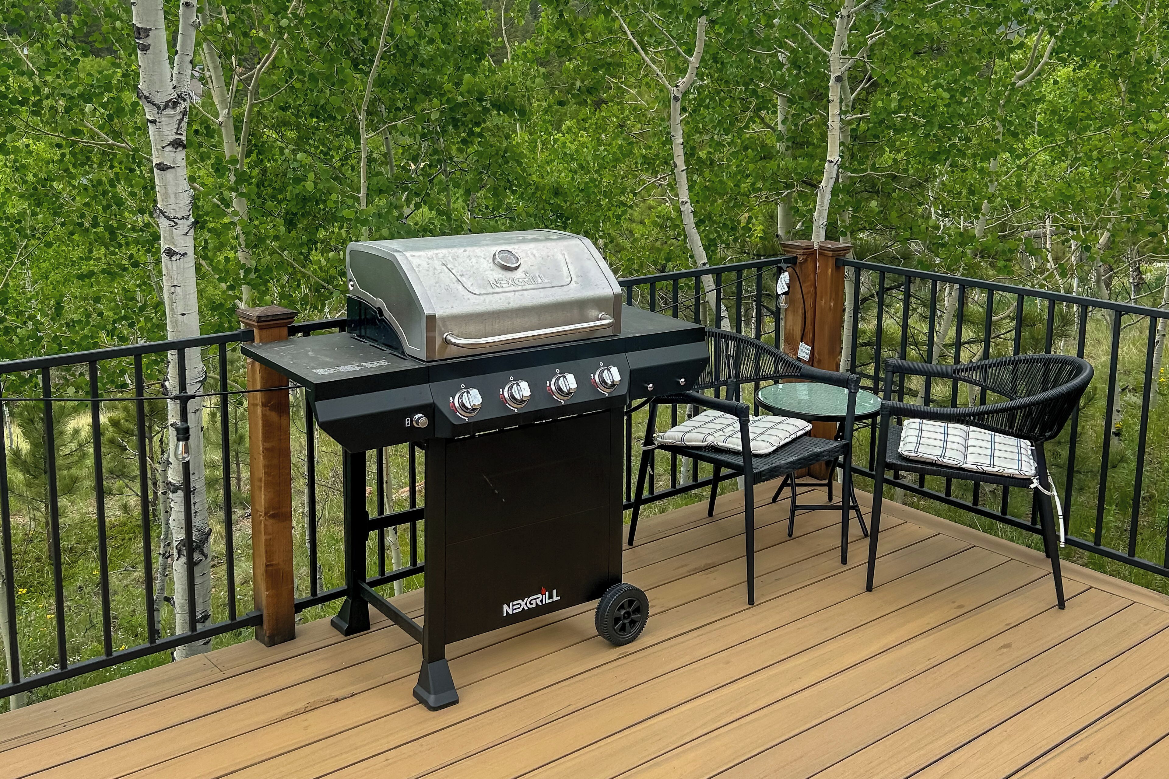 Un barbecue NexGrill sur une terrasse en bois, avec une table et des chaises, entouré d’une rambarde en métal et d’arbres.