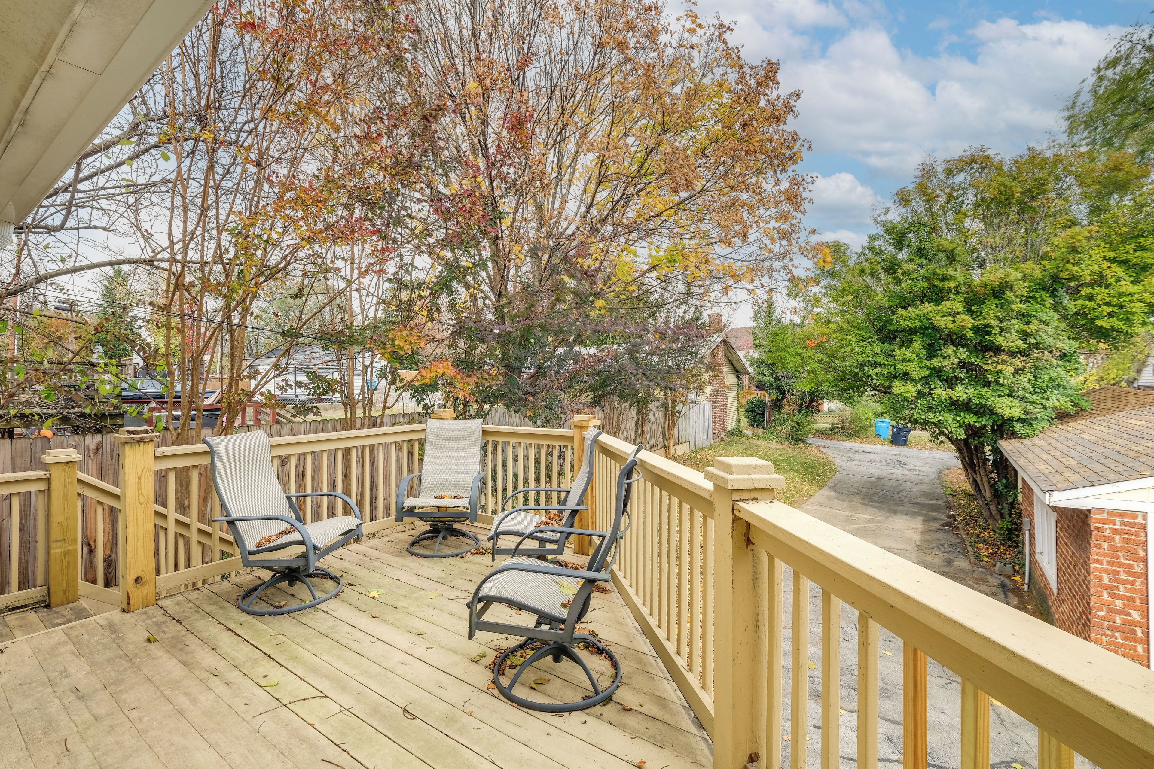A wooden deck with two chairs and a table, surrounded by a wooden railing, with trees and a house in the background.