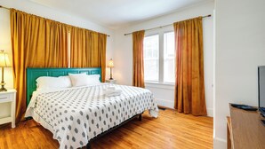 Ein Hotelzimmer mit Holzboden, einem türkisblauen Kopfteil, einem Bett mit weißen Leinen und zwei mit Vorhängen versehenen Fenstern.