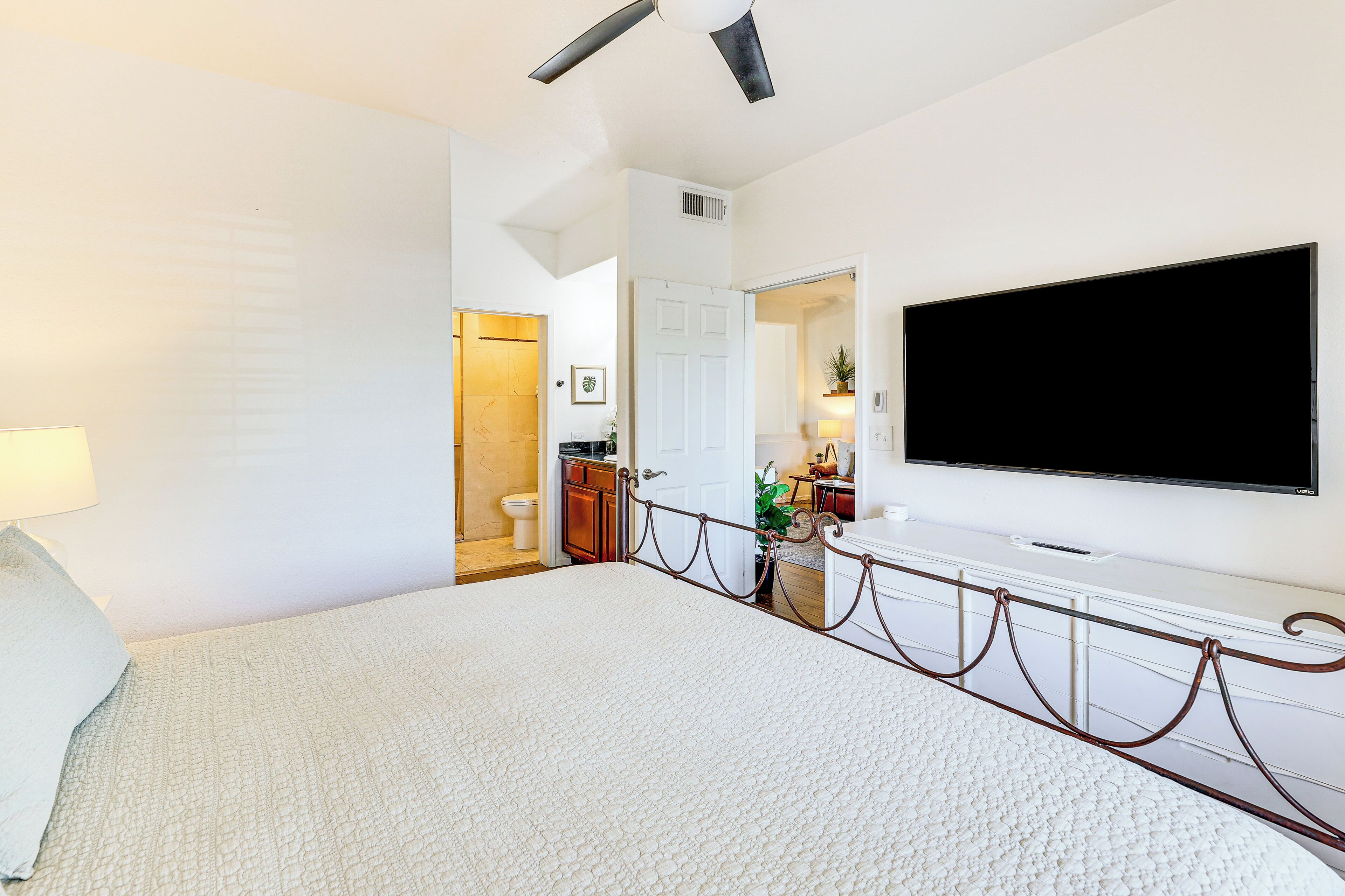 Una habitación de hotel con una cama grande, un televisor de pantalla plana montado en la pared, un baño con inodoro y lavamanos, y una mesita con una planta.