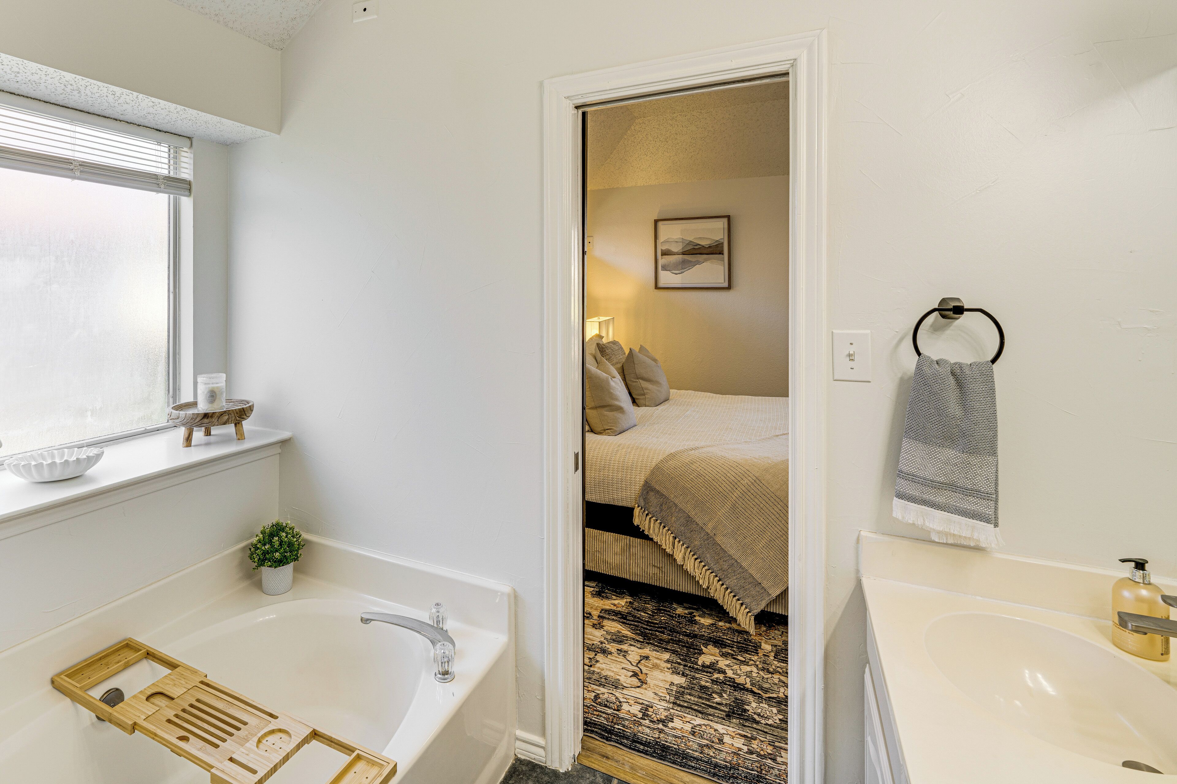 A bathroom with a bathtub, a sink, a window, a small plant, and a door leading to a bedroom.