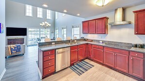 A spacious kitchen with a built-in dishwasher, red cabinets, and a large island with a sink. There is a dining area with chairs and a TV in the background. - St Joseph River, Lake Michigan Access: Luxury Home (Saint Joseph)