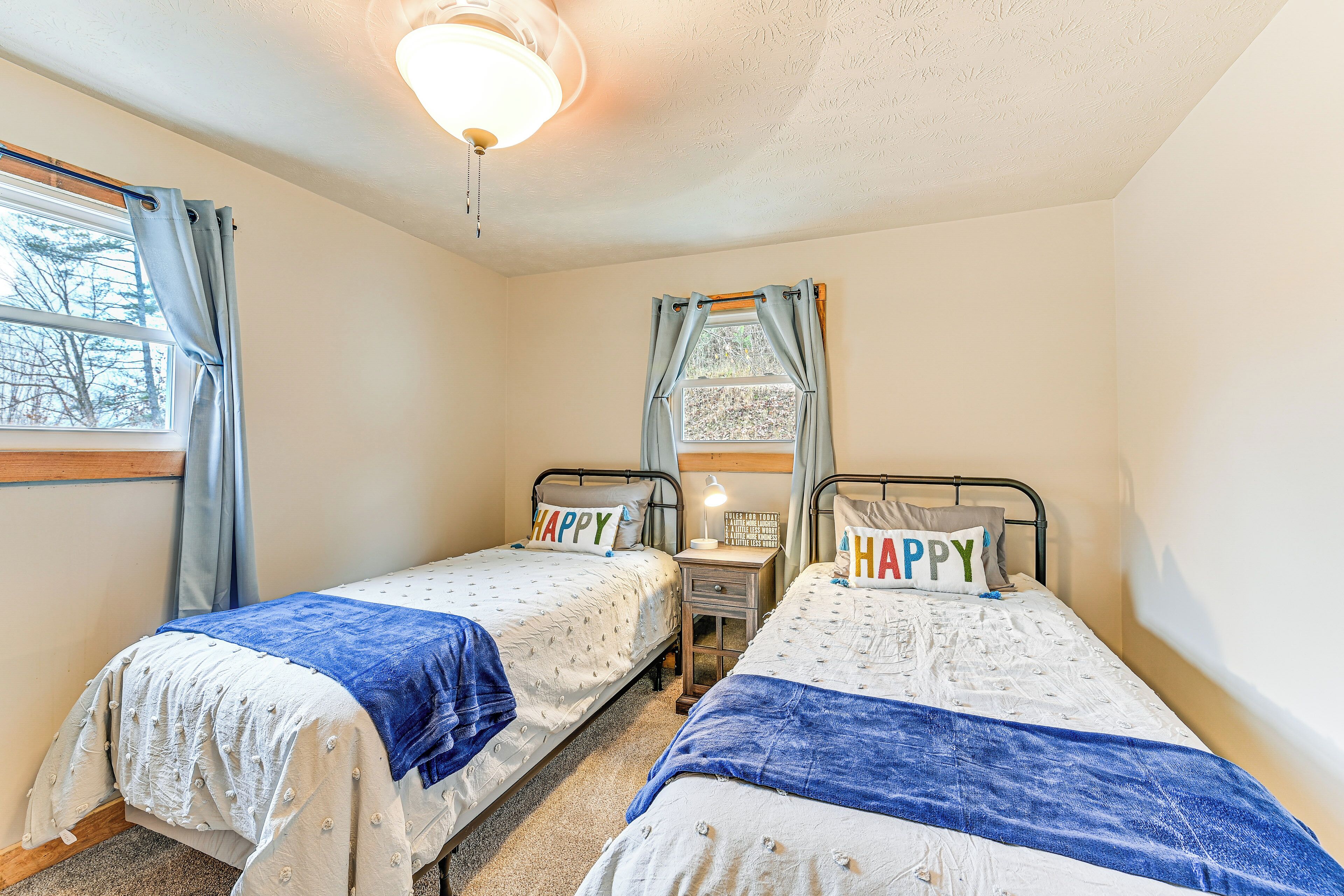 Une chambre avec deux lits simples, chacun agrémenté d’un coussin orné du mot « HAPPY », une petite table de chevet avec une lampe et une fenêtre avec des rideaux.