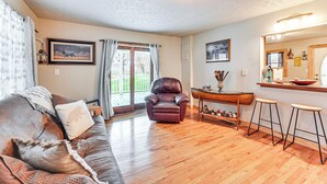 A cozy living room with a leather sofa, a leather armchair, and a wooden coffee table. There is a large window with curtains, a chandelier, and a mirror above the coffee table. - On 220 Acres: Home w/ Trails in West Liberty! (West Liberty)