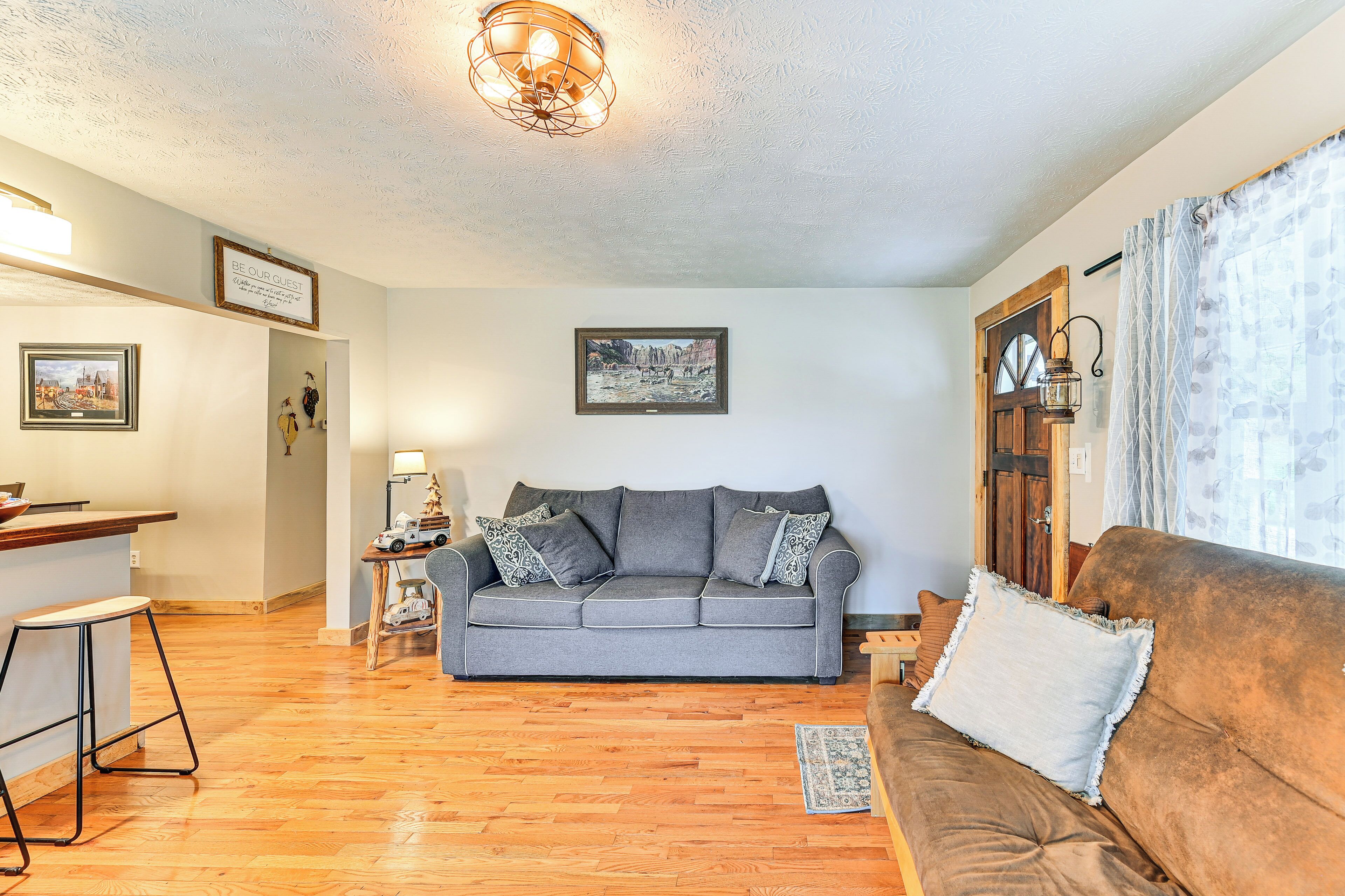 Ein Wohnzimmer mit grauem Sofa, braunem Sofa, Holzboden, gerahmtem Bild an der Wand und kleinem Tisch mit Lampe.