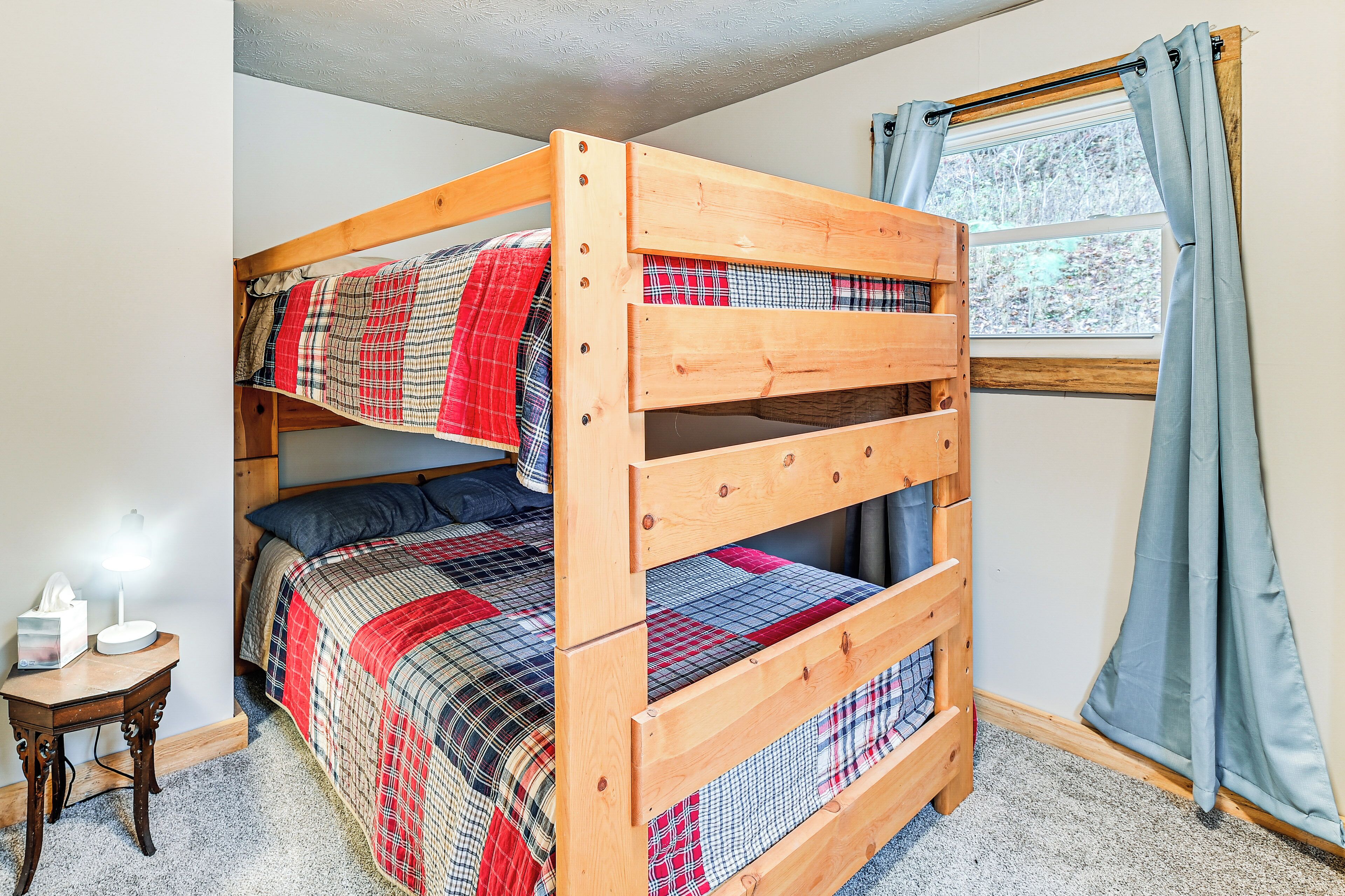 A bunk bed with a plaid bedspread, a bedside table with a lamp, and a window with curtains.