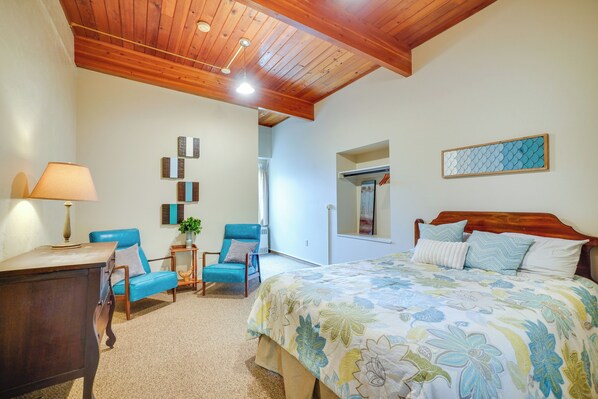 A bedroom with a wooden ceiling, a bed with floral bedding, a desk with a lamp, and two blue chairs. - Grand Rural Wisconsin Retreat: Full Indoor Gym (Mount Calvary)