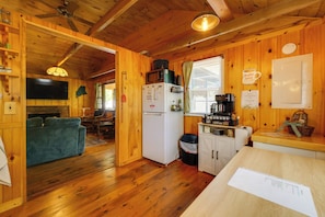 A wooden cabin interior with a kitchen area, a dining table, and a television mounted on the wall. - 'knotty Kabin' Rustic Waterboro Cabin By Lake (Waterboro)