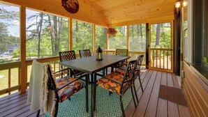 A covered outdoor deck with a table and chairs, large windows, and a view of trees.