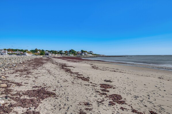 A beach with a rocky shoreline, clear blue sky, and houses in the background. - Steps to North Hampton Beach: Cottage w/ Deck! (North Hampton)