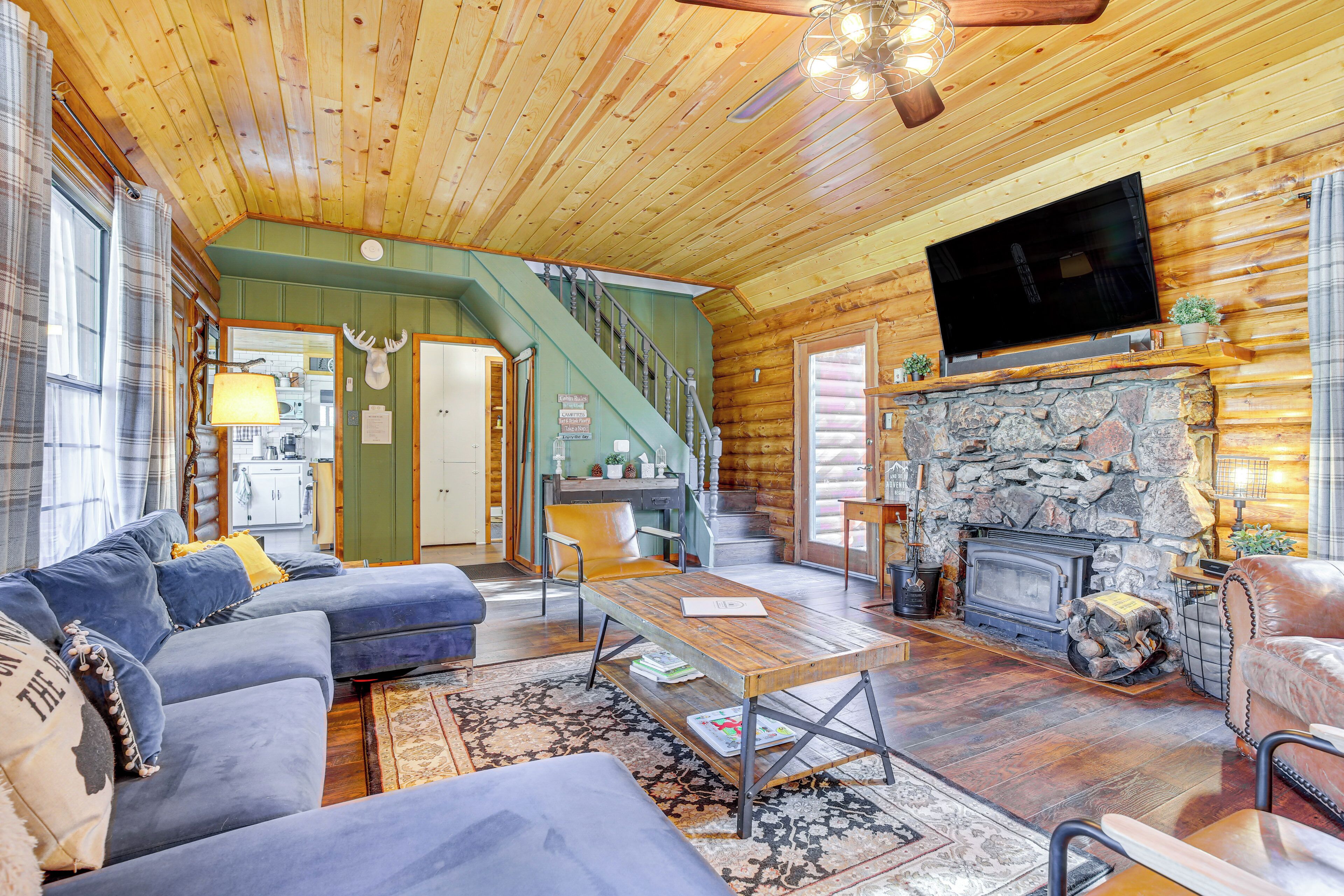 A cozy log cabin interior with a living room, a stone fireplace, a television mounted above it, and a staircase leading to a kitchen area.