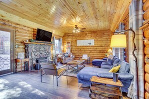 Interior de uma cabana de tronco com lareira de pedra, móveis de madeira e televisão fixada na parede.
