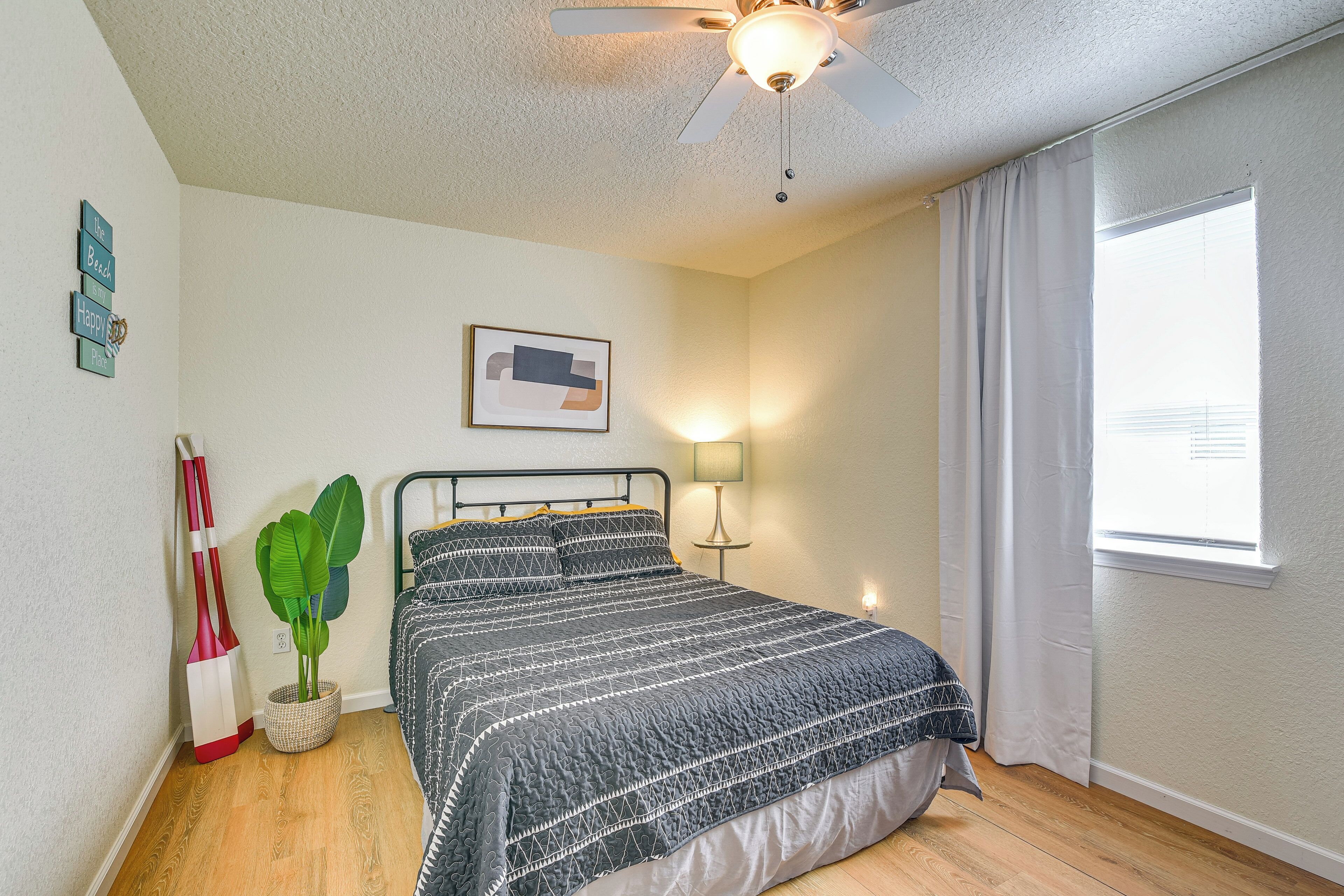 A bedroom with a bed, a ceiling fan, a window with curtains, a wall-mounted lamp, and a framed picture.