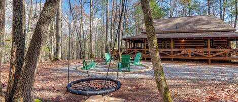 A wooden cabin with a swing set and outdoor seating area surrounded by trees.