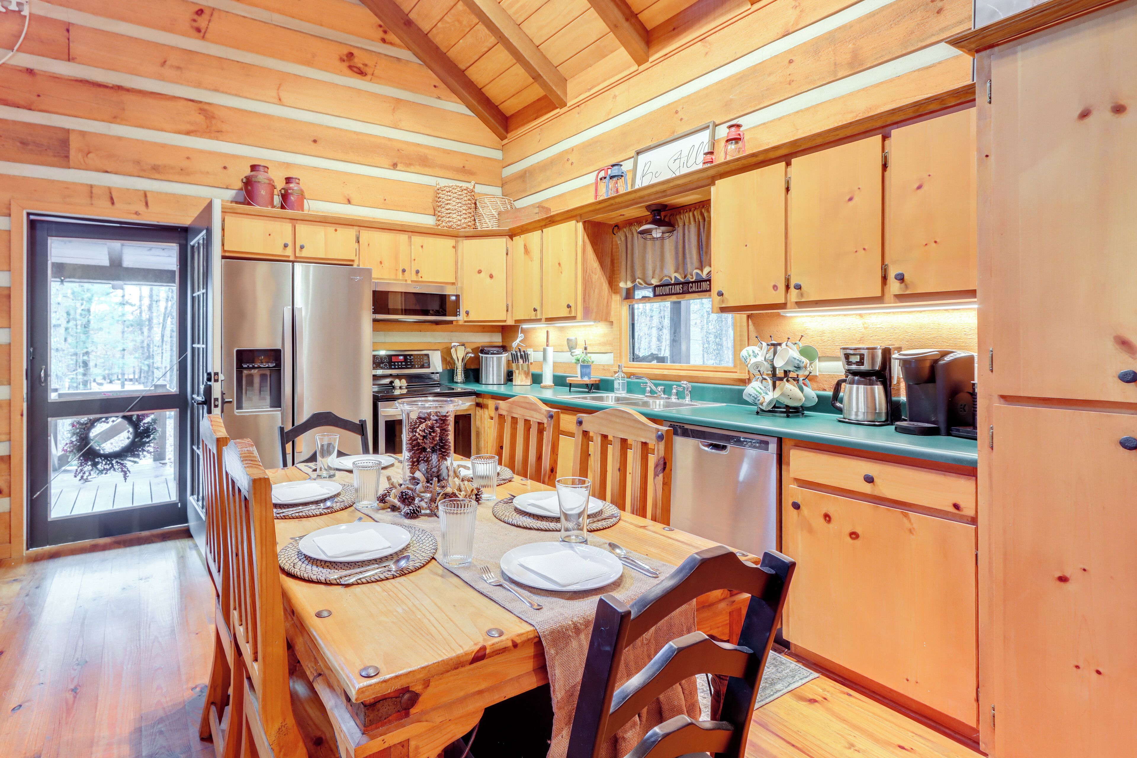 A wooden cabin interior with a dining table set for four, wooden cabinets, a refrigerator, and a window with a wreath.