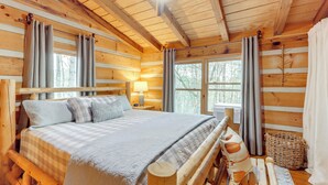 A log cabin bedroom with a wooden bed, a nightstand, a lamp, and a window with curtains. - Ridgetop Cabin in Blue Ridge! Wraparound Porch! (Blue Ridge)