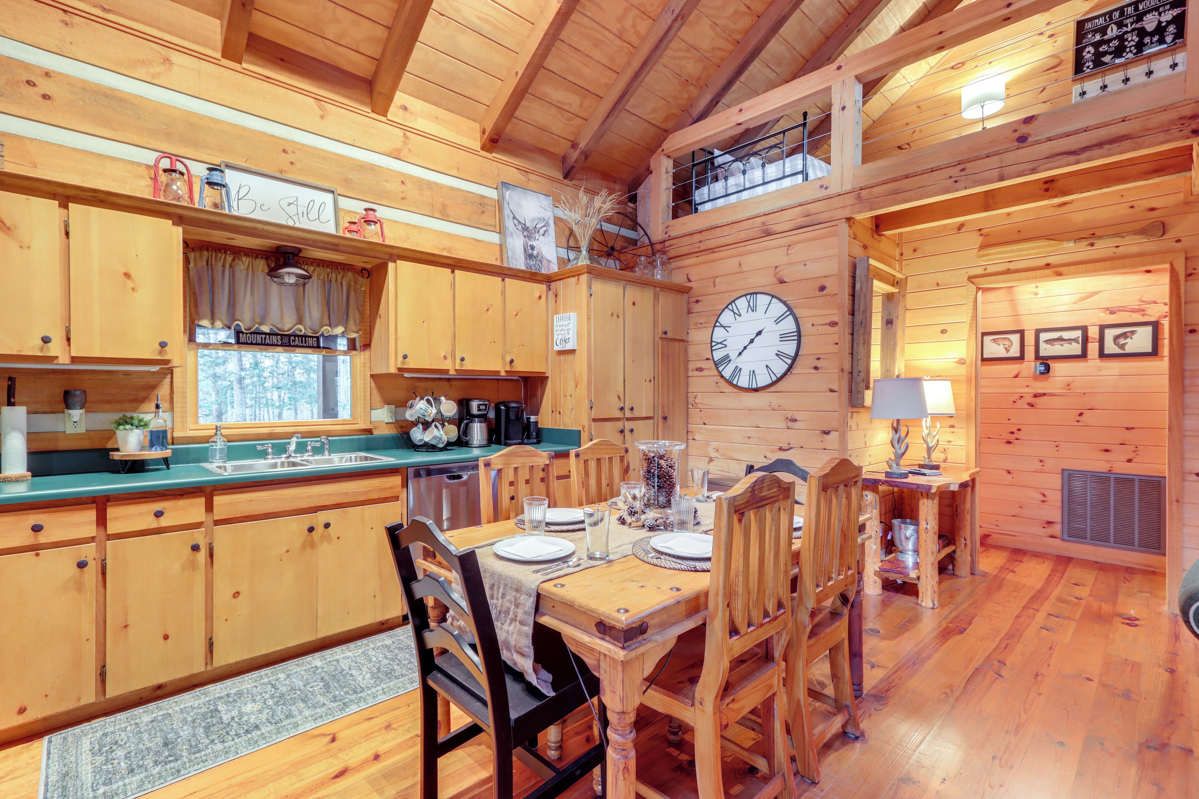 A wooden cabin interior with a dining table set for four, wooden cabinets, a clock, and a sign reading "Mountains Calling".