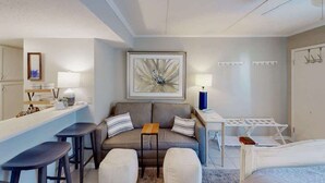 Une chambre d’hôtel moderne comprenant un canapé, deux tabourets de bar, une petite table, un lit, un bureau avec une lampe, une étagère murale et un grand tableau abstrait.