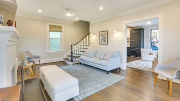 A spacious living room with a light blue sofa, a white ottoman, and a patterned rug. A staircase with a wooden railing leads to another room.
