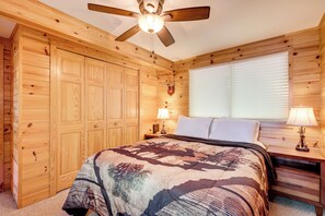 Quarto com painéis de madeira, uma cama, duas mesas de cabeceira, um ventilador de teto e uma janela com persianas.
