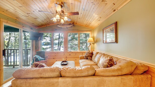 A cozy living room with a wooden ceiling, a large window, and a sofa. - Dock + Fire Pit: Sweet Serenity on Amber Lake (Lac du Flambeau)