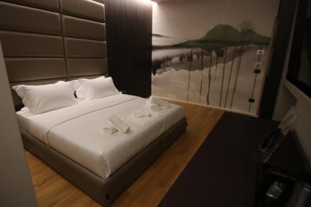 A hotel room with a bed, a headboard, a wall with a scenic mural, and a small table with a lamp.