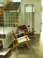 Un interior moderno con una escalera de cristal, sillas de madera y una lámpara de pie.