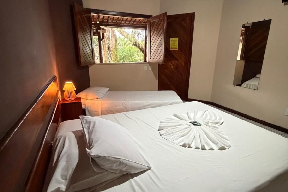 A hotel room with two beds, a nightstand with a lamp, a window with curtains, and a wooden ceiling.
