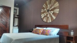 A bedroom with a bed, a wooden headboard, a wall clock, and a small shelf. - POUSADA PORTAL DO SOL  (Itacaré)
