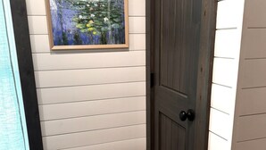 A room with white paneled walls, a dark wooden door, and a framed painting of a pond with flowers. - Cozy 2 Bedroom Cottage in the Woods, Near Petoskey, Golf Courses and Ski Resorts (Petoskey)