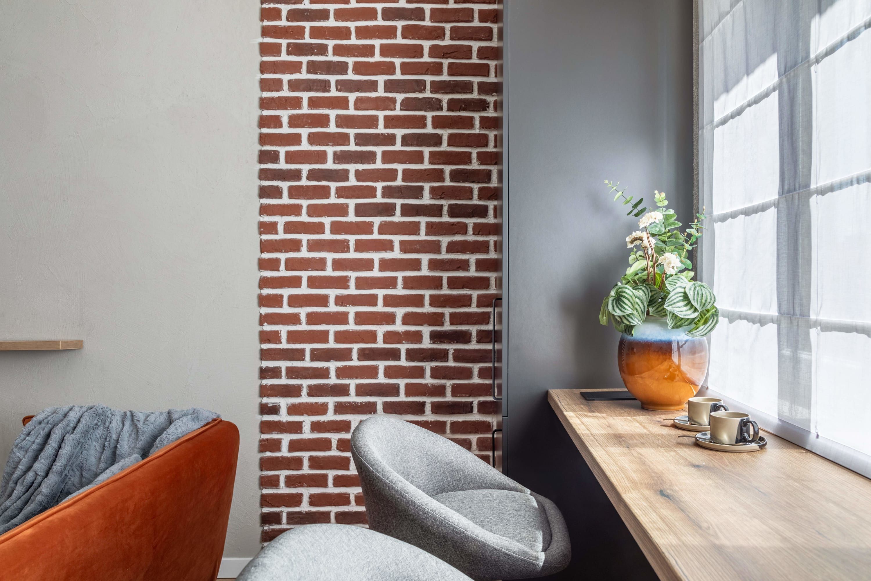 A corner of a room with a brick wall, a window with blinds, a wooden table with two cups, and a potted plant.