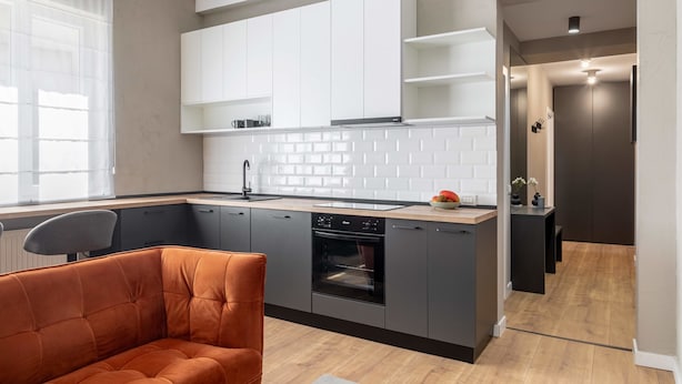A modern kitchen with white cabinets, a dishwasher, and a built-in oven. There is a window with sheer curtains and a chair in the foreground. - Holt Rossetti Residences Exclusive Urban Lofts (Bucharest)