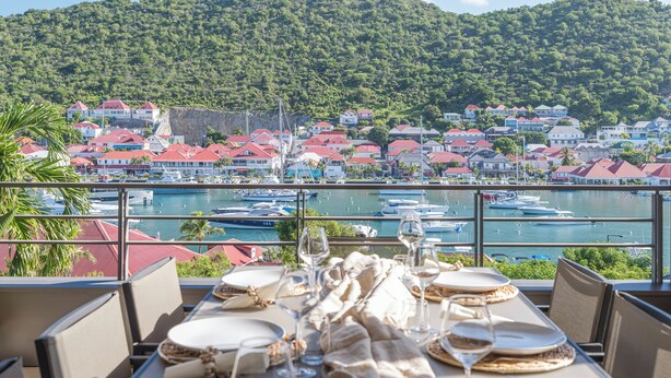A dining table set with plates, glasses, and cutlery, overlooking a marina with boats and a coastal town with red-roofed buildings. - Beautiful 2-bedroom apartment in Gustavia with harbor view in Saint Barthélémy (Gustavia)